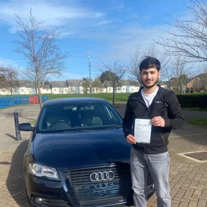 Learner in a black jacket holding a pass certificate beside a dark Audi