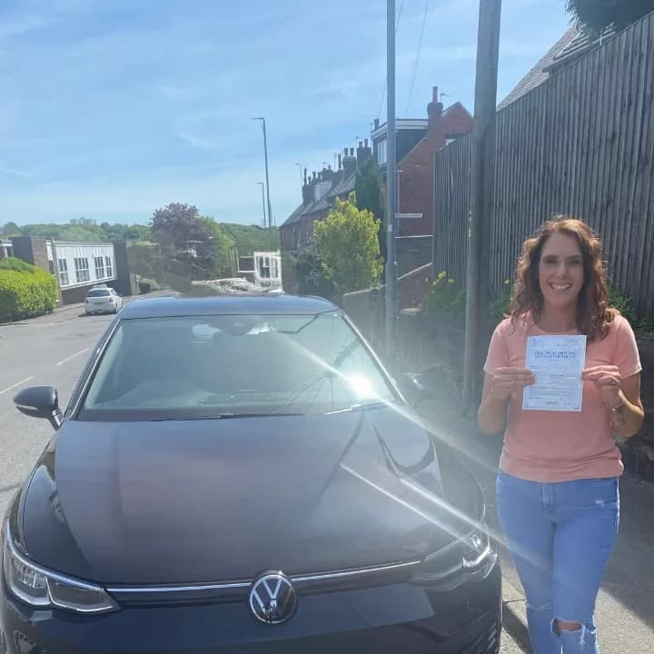 Learner in a pink top holding a pass certificate beside the black VW Golf